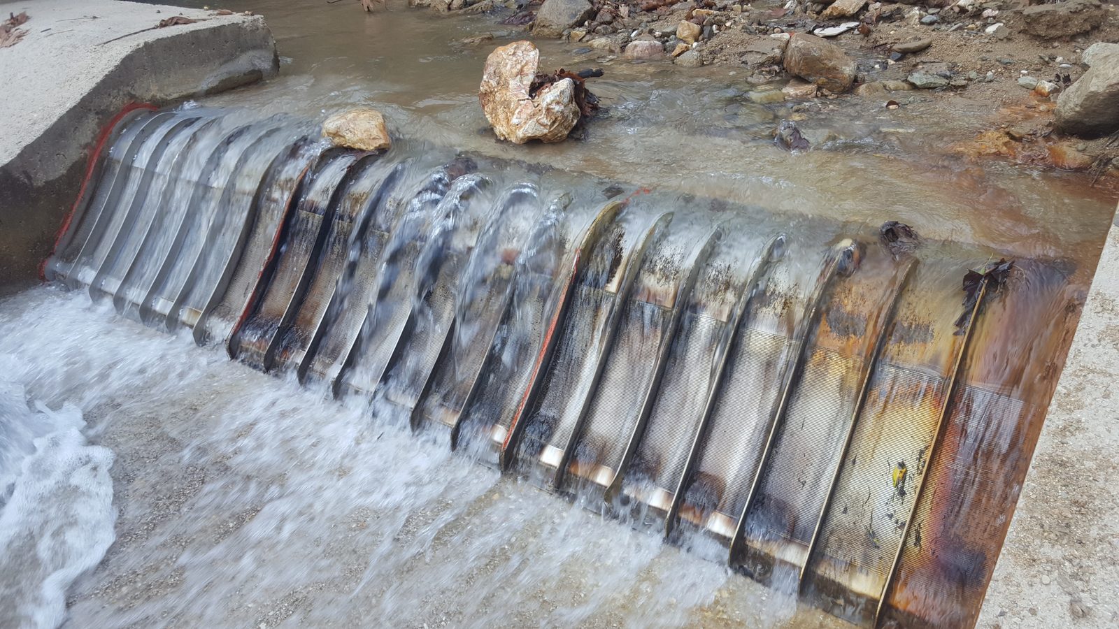 Dieselbe Wasserfassung Ende August 2018, nach dem Hochwasser: das Becken hinter dem Wehr bis zur Wehrkrone mit Steinen und Sand gefüllt, Blöcke liegen auf dem Rechen, Wasser fließt weiterhin durch den Keildraht