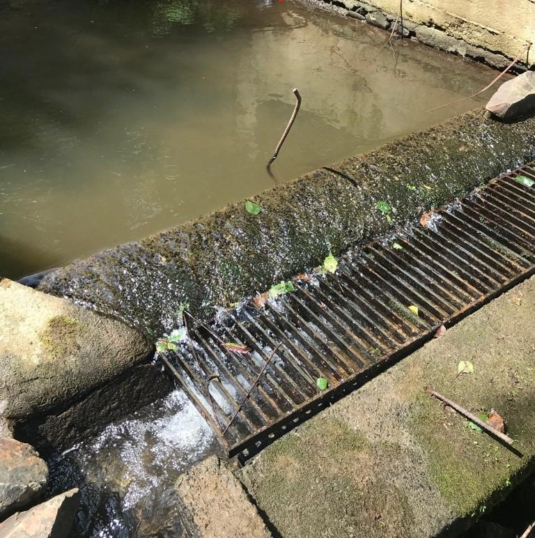 Çarşıbaşı, Yavuz intake before the retrofit: a rusted flat bar grate on the weir crest, holding leaves and debris, with murky water pooled behind the weir